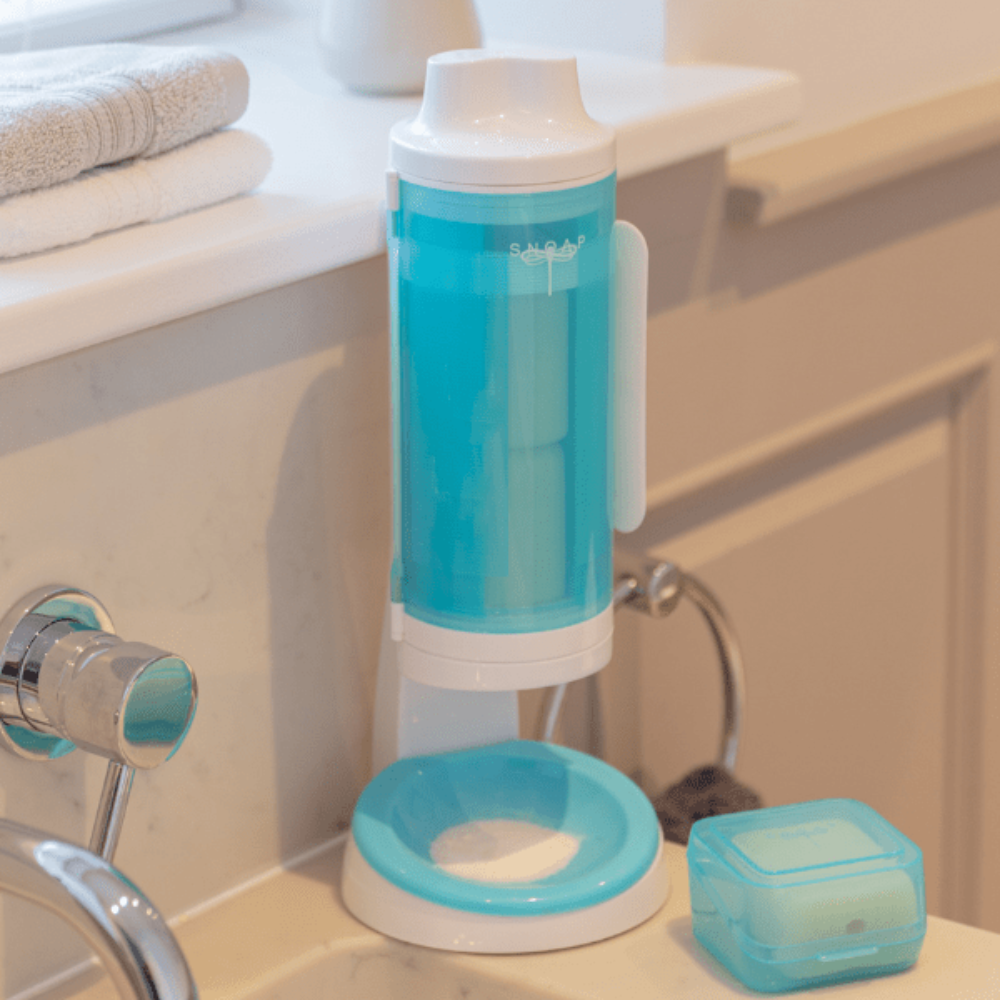 SNOAP Dispenser in white & SNOAP blue, counter mounted by bathroom sink with soap box next to it.
